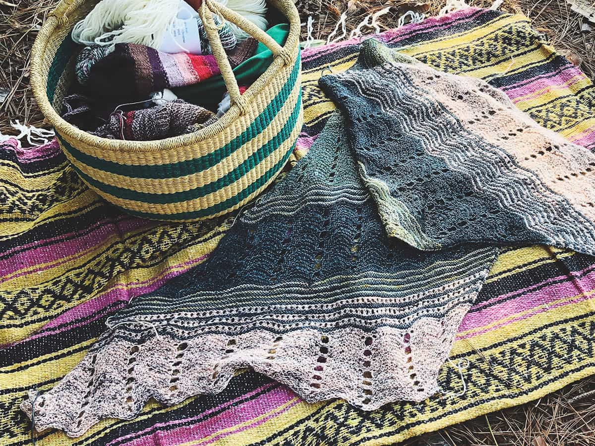 Image of my Dirty Lace hand knit wrap on a Mexican blanket beneath pine trees.