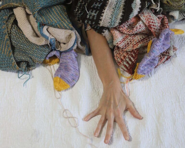 Image of me reaching out from under a massive pile of knits that need their ends woven in.