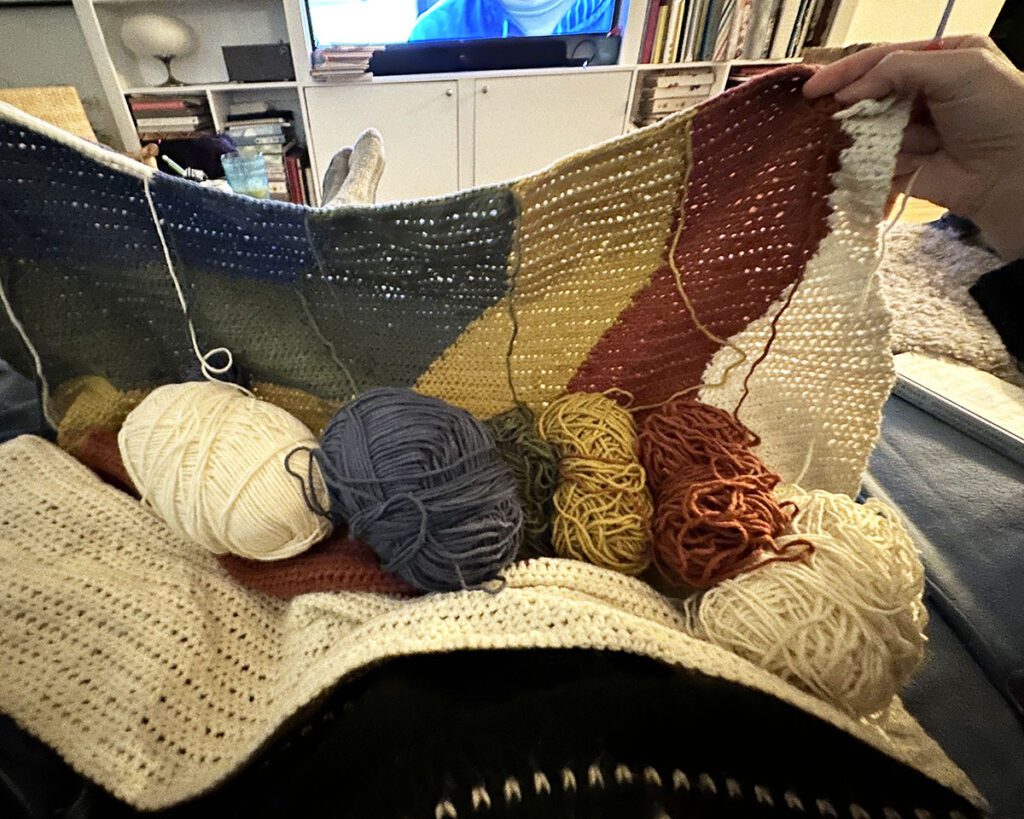 Image of me sitting with an intarsia crochet blanket that has 6 balls of yarn hanging from it. It covers me.