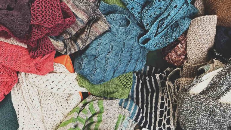 Image of a big pile of summer sweaters on the bed beside me from where I review all of the yarns for you in this vlog.